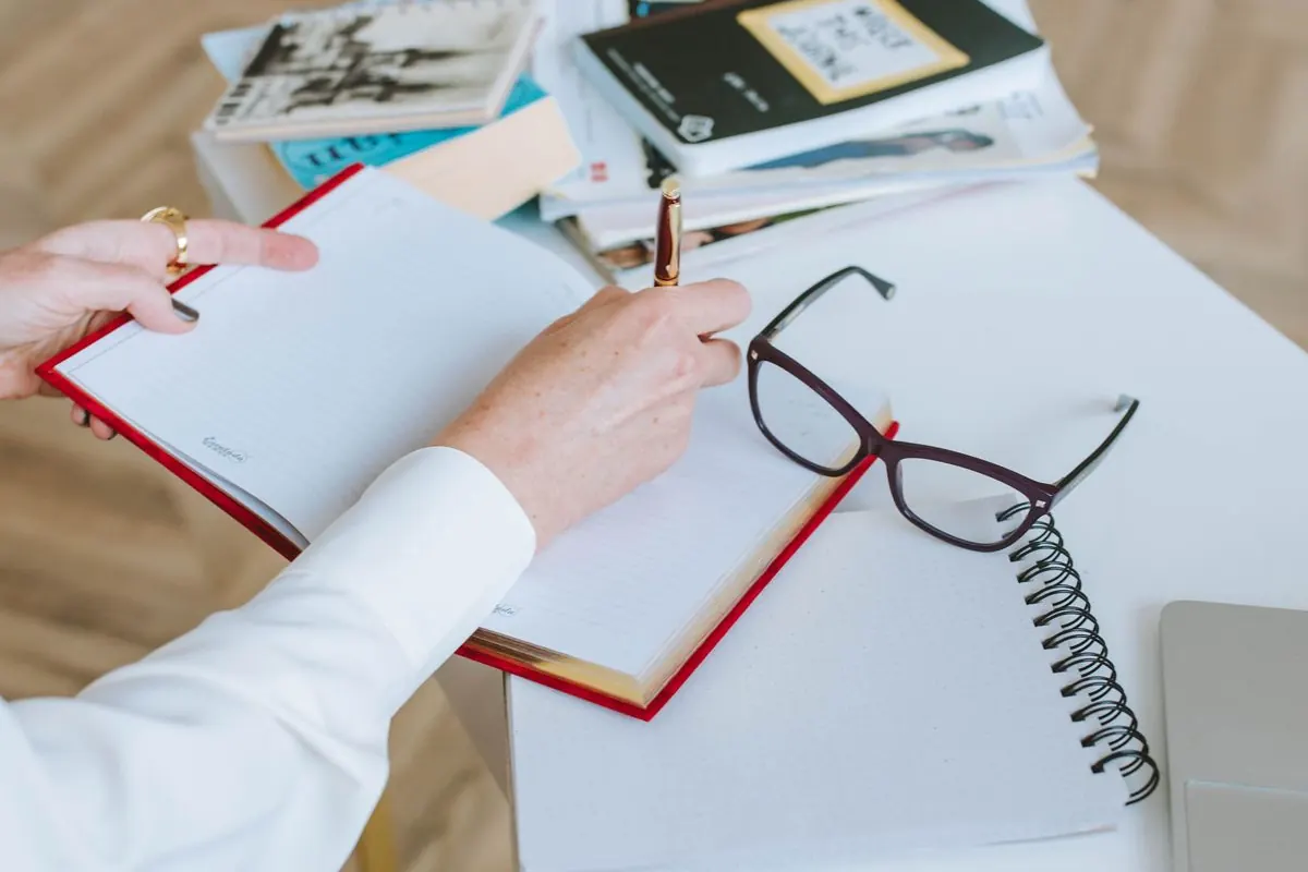 Journaling at a desk with a notebook, pen, glasses, and a stack of books