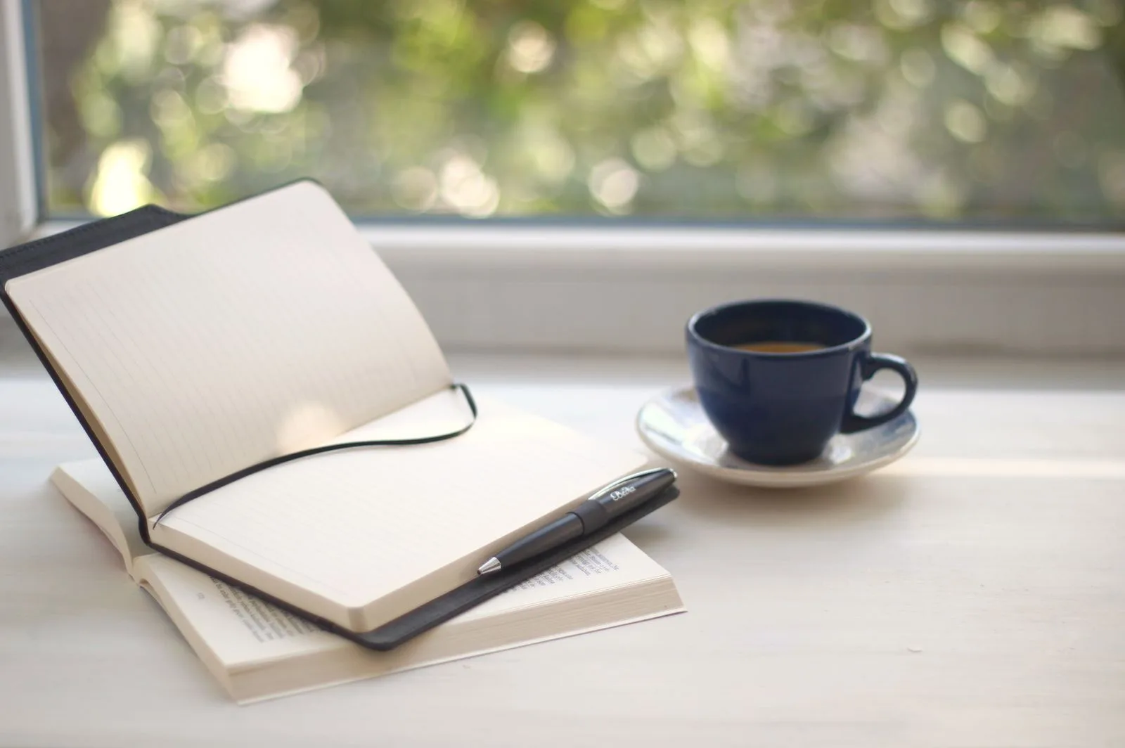 An open notebook and pen beside a cup of coffee on a windowsill in soft morning light