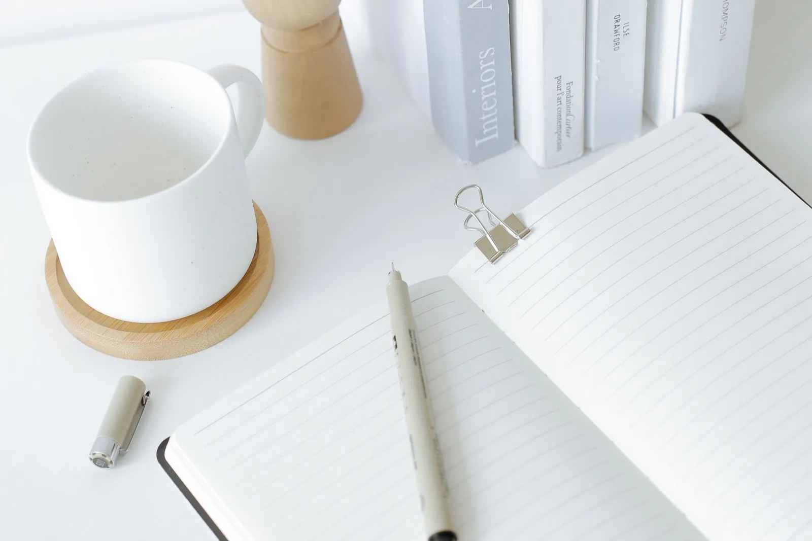 An open notebook and pen on a white desk beside a stack of books and a mug of coffee
